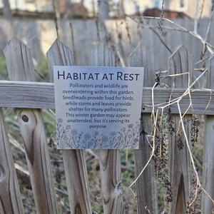 May include: A rectangular metal sign with the words "HABITAT AT REST" in black text is attached to a weathered wooden fence. The sign's text describes the garden's role in supporting pollinators and wildlife during winter. Dried plants are visible.