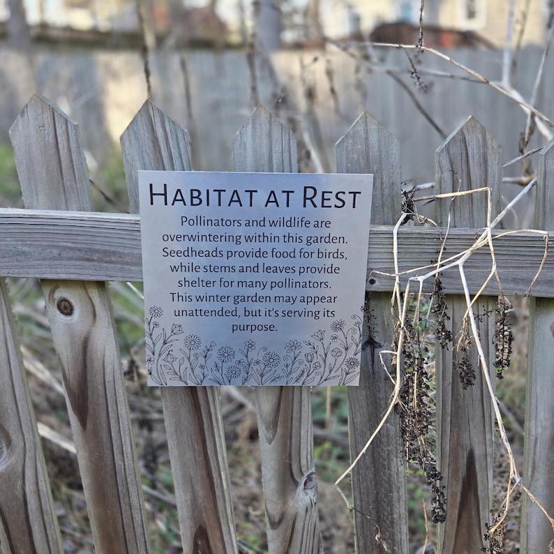 May include: A rectangular metal sign with the words "HABITAT AT REST" in black text is attached to a weathered wooden fence. The sign's text describes the garden's role in supporting pollinators and wildlife during winter. Dried plants are visible.