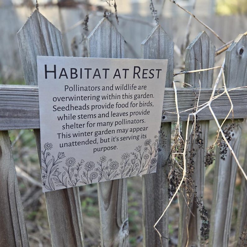 May include: A sign with the text "HABITAT AT REST" is attached to a wooden fence. The sign describes how pollinators and wildlife overwinter in the garden. The sign has a floral border.