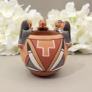 Puede incluir: Una pequeña vasija de barro color terracota con tres figuras esculpidas unidas al borde. El exterior está decorado con patrones geométricos en blanco, negro y beige, incluyendo un motivo central en forma de T y detalles triangulares.
