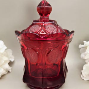 May include: A deep red glass jar with a matching lid, featuring a scalloped rim and embossed circular medallions on the body and lid. The glass is translucent and vibrant, showcasing a repeating pattern of detailed circular motifs across the surface.