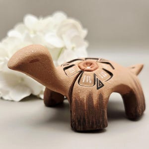 May include: A terracotta turtle figurine with a floral design on its back. The turtle has a light brown body with darker brown accents on its legs and tail. The floral design is in white and black.