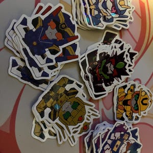 May include: A collection of die-cut stickers featuring various robot characters in different colour schemes including blue, yellow, black, and green. Each sticker has a white border and depicts a stylised, chibi-inspired mechanical figure.