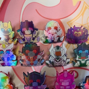 Könnte beinhalten: Eine Sammlung farbenfroher, holografischer Roboter-Charakter-Anstecknadeln. Jede Anstecknadel hat ein einzigartiges Design mit leuchtenden Farbtönen von Gelb, Lila, Blau und Rosa. Die Anstecknadeln sind auf einer rosa-weiß gemusterten Oberfläche angeordnet.