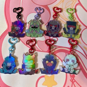 Op de afbeelding: Een verzameling van acht sleutelhangers met robotthema's en hartvormige sluitingen. De sleutelhangers hebben verschillende kleuren, waaronder blauw, paars, groen en rood, met een holografisch effect. Elke sleutelhanger heeft een uniek robotontwerp.