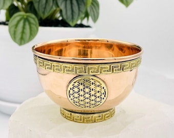 May include: A small copper bowl with a gold trim and a flower of life design in the center. The bowl is sitting on a white surface.