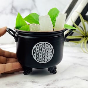 May include: Black cauldron with three legs and a white flower of life design. The cauldron is filled with selenite sticks and green plant leaves.