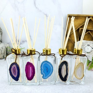 May include: Five glass bottles with gold lids and wooden sticks, each containing a different colored agate slice. The bottles are arranged in a row on a white surface.