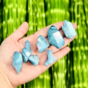May include: A collection of eight blue iridescent quartz crystals, also known as angel aura quartz, in various sizes and shapes. The crystals are arranged on a hand, with the background blurred.