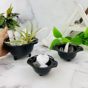 Puede incluir: Tres ollas negras en forma de caldero con asas. La olla más grande contiene una planta verde y cristales blancos. La olla del medio contiene un cristal blanco en forma de corazón y otros cristales. La olla más pequeña contiene cristales blancos.