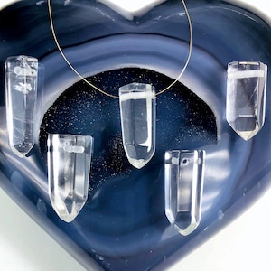 May include: Five clear quartz crystal points with a hole drilled through the top, displayed on a blue heart-shaped agate. The crystals are arranged in a row, with the top one hanging from a thin gold chain.