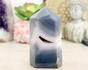 Pode incluir: Uma ponta de cristal de ágata cinza e branca com uma forma natural irregular. O cristal tem um padrão de bandas sobrepostas com um acabamento liso e polido.