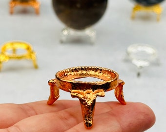 May include: A gold-colored metal stand with three legs and a round top, designed to hold a crystal ball or other small object. The stand is made of a textured metal and has a slightly worn look.