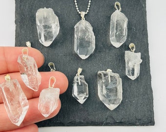 May include: Eight clear quartz crystal pendants with silver bail settings. The pendants are various sizes and shapes, and are displayed on a black slate surface.