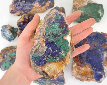 May include: A collection of raw azurite mineral specimens. The stones display a mix of deep blue, green, and brown hues. The azurite is held in a hand, with other specimens scattered on a white surface.