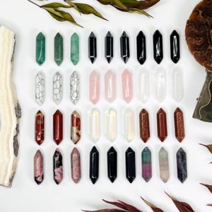 May include: A collection of 36 double-terminated crystal points in various colors and patterns, including green, black, white, pink, red, and brown. The crystals are arranged in four rows of nine, on a white surface.