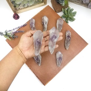 May include: Seven amethyst crystal points with a purple hue, arranged on a brown surface. The crystals are pointed and have a natural, raw look.