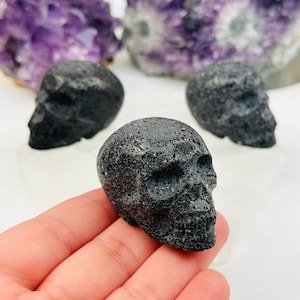 Puede incluir: Tres esculturas de cráneos de obsidiana negra. Los cráneos son lisos y tienen una superficie rugosa y texturizada.