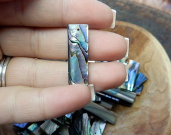 May include: A close-up of a pile of iridescent abalone shell pieces. The pieces are rectangular and have a variety of colors, including blue, green, and purple. The pieces are arranged in a pile on a wooden surface.