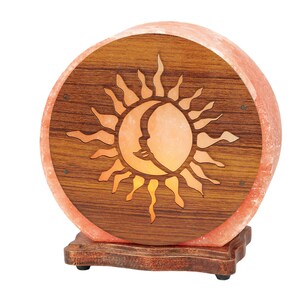 K&ouml;nnte beinhalten: Eine runde Himalaya-Salzlampe mit einem Holzsockel und einem geschnitzten Sonnen- und Mondmotiv. Die Lampe ist eingeschaltet und leuchtet orange.