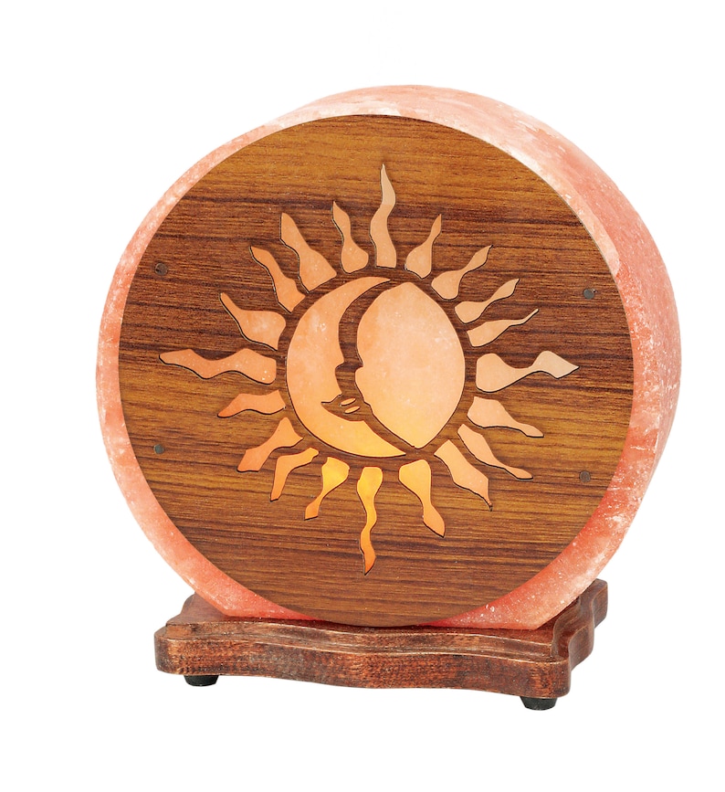 K&ouml;nnte beinhalten: Eine runde Himalaya-Salzlampe mit einem Holzsockel und einem geschnitzten Sonnen- und Mondmotiv. Die Lampe ist eingeschaltet und leuchtet orange.