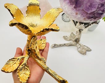 May include: A gold-coloured metal flower-shaped decorative stand with a stem and leaves. A second silver-coloured metal stand holds a large purple amethyst crystal sphere. The stands are on a white surface.