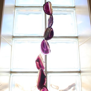 May include: A purple agate crystal mobile hanging from a white ring in front of a window. The crystals are suspended from a clear string and are arranged in a vertical line.