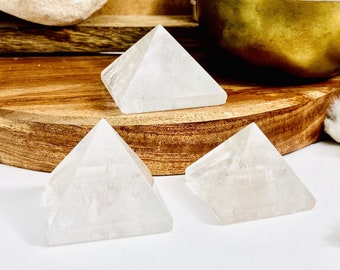 May include: Three clear quartz crystal pyramids on a wooden surface. The pyramids are smooth and polished, with a slight sparkle.