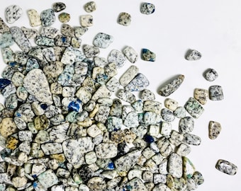 May include: A collection of tumbled K2 stones, also known as azurite in granite, scattered on a white surface. The stones vary in size and shape, displaying a mix of white, gray, blue, and black speckles and patches. The overall aesthetic is natural and earthy.