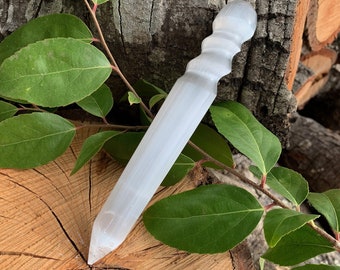 Puede incluir: Varita de selenita blanca con una punta redondeada y una superficie texturizada. La varita mide aproximadamente 18 cm de largo y está apoyada sobre un trozo de madera.