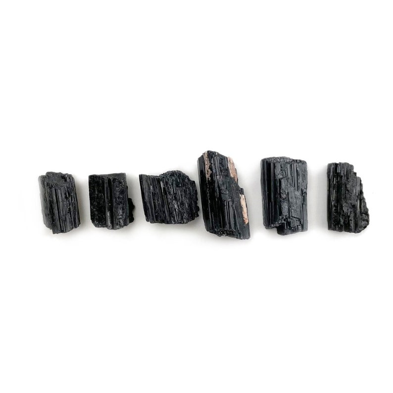 May include: Six black tourmaline crystals, also known as schorl, are arranged in a row on a white background. The crystals are raw and unpolished, with a natural, rough texture.