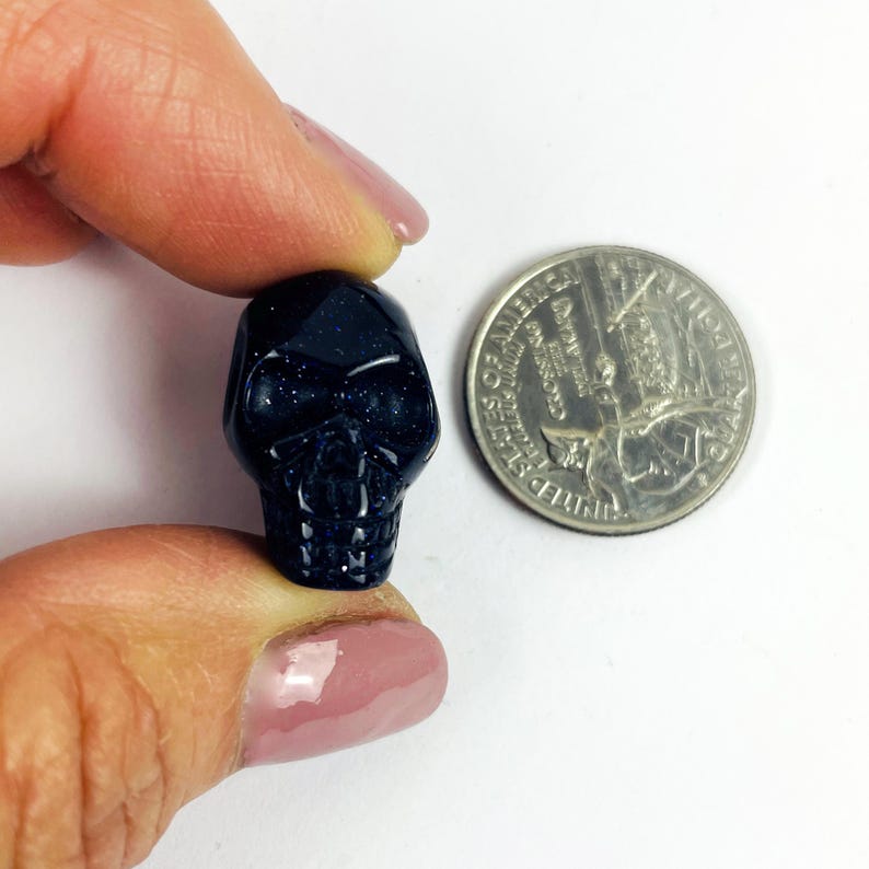 May include: Skull Bead-- Purple Goldstone Skull Bead on hand next to quarter on white surface for reference.