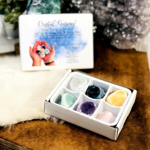 Könnte beinhalten: Eine weiße Box mit sechs Fächern, die verschiedene farbige Kristallsteine enthalten. Die Box ist mit "Crystal Greeting!" beschriftet und zeigt eine Aquarell-Illustration von Händen, die Kristalle halten.
