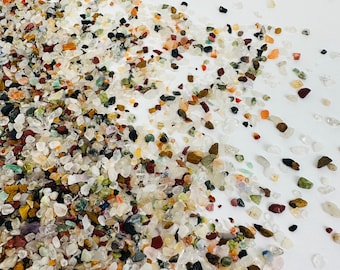 May include: A close-up of a variety of small, colorful stones and crystals. The stones are scattered on a white surface and range in color from white to red, brown, and green.