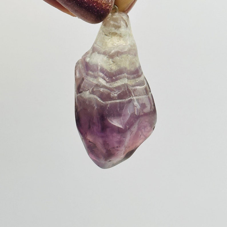 May include: A purple amethyst crystal pendant with a clear, white, and purple layered pattern. The crystal is shaped like a teardrop and has a smooth, polished surface.
