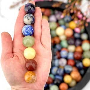 May include: A hand holding a line of seven chakra healing crystal spheres. The spheres are amethyst, sodalite, lapis lazuli, aventurine, citrine, carnelian, and tiger's eye. The spheres are arranged in a rainbow order, starting with purple and ending with yellow. The background is a blurred image of a bowl filled with many colorful crystal spheres.