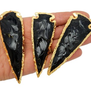 May include: Three black obsidian arrowheads with gold-plated edges. The arrowheads are displayed on a hand.