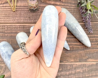 May include: A light blue crystal wand, also known as a yoni egg, weighing between 100 and 200 grams. The wand is smooth and polished, with a pointed tip and a rounded base.