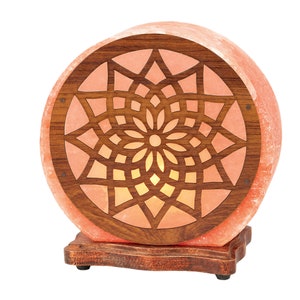 K&ouml;nnte beinhalten: Eine runde Himalaya-Salzlampe mit einem Holzsockel und einem geschnitzten geometrischen Muster. Die Lampe ist beleuchtet und strahlt ein warmes Licht aus.