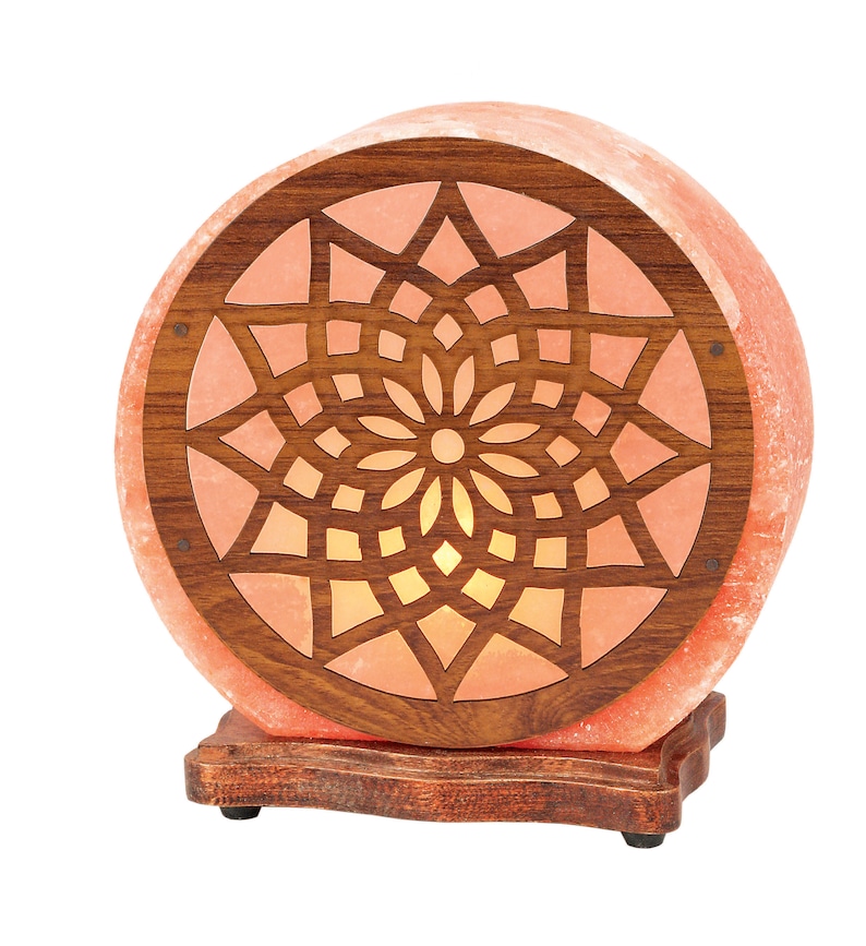K&ouml;nnte beinhalten: Eine runde Himalaya-Salzlampe mit einem Holzsockel und einem geschnitzten geometrischen Muster. Die Lampe ist beleuchtet und strahlt ein warmes Licht aus.