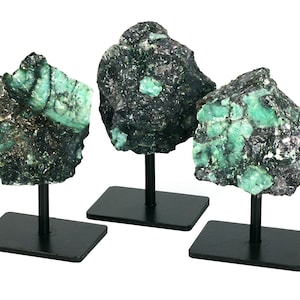 May include: Three emerald green gemstone clusters on black metal stands. The clusters are rough and have a dark green matrix.