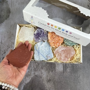May include: A box set of raw crystals in various colors, including clear, purple, orange, green, blue, and pink. The crystals are displayed on straw inside a cardboard box with the text "Crystals for the Mind, Body and Soul!"