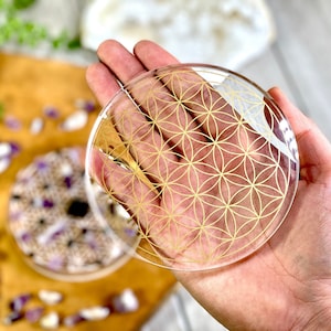 May include: A clear glass coaster with a gold flower of life design. The coaster is round and has a smooth surface.