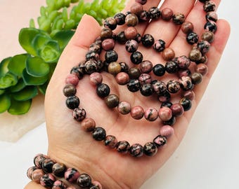 May include: A collection of beaded bracelets featuring pink, brown, and black spherical beads. The bracelets are held in a hand, highlighting the different colours and patterns of the stones. The beads appear to be approximately 0.8 cm in diameter.