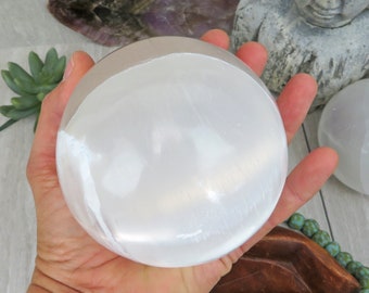 May include: A white selenite sphere, also known as a selenite ball, is held in a hand. The sphere is smooth and polished, with a slightly translucent appearance.