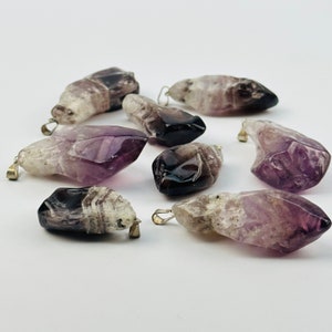 May include: A collection of eight amethyst crystal pendants with silver-toned metal bail loops. The crystals are a mix of purple, white, and black.