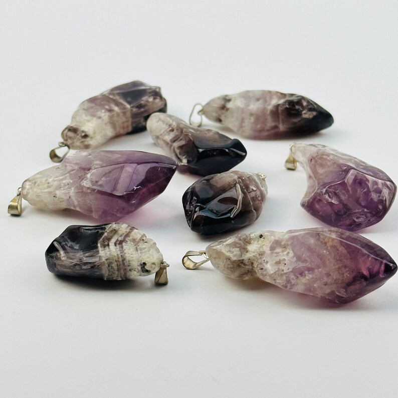 May include: A collection of eight amethyst crystal pendants with silver-toned metal bail loops. The crystals are a mix of purple, white, and black.