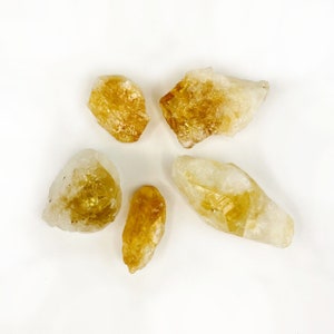 Puede incluir: Cinco cristales de citrino sin pulir, de color amarillo, de diferentes tama&ntilde;os y formas. Los cristales est&aacute;n sobre un fondo blanco.