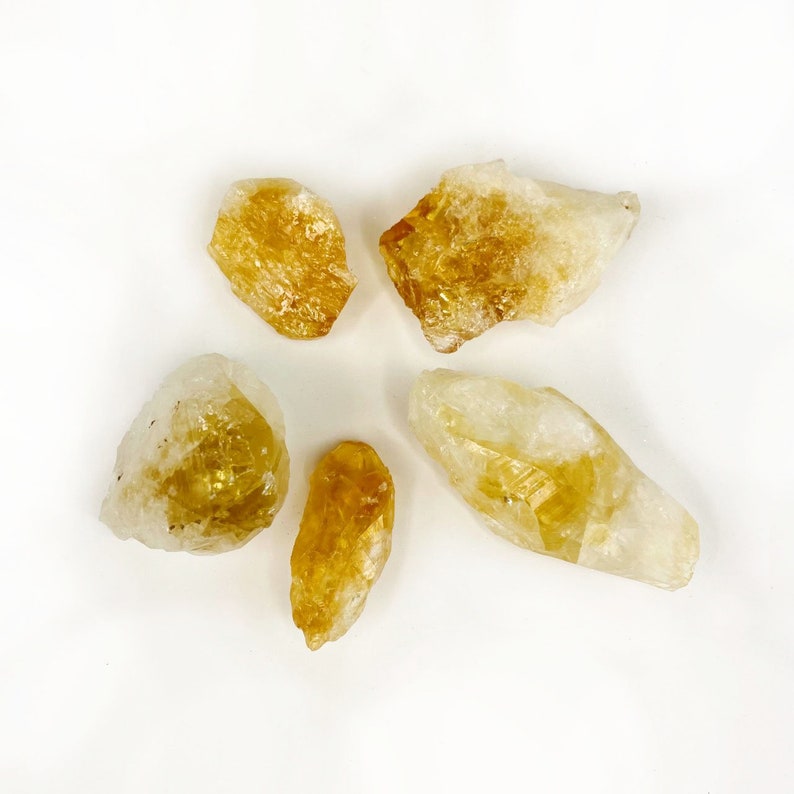 Puede incluir: Cinco cristales de citrino sin pulir, de color amarillo, de diferentes tama&ntilde;os y formas. Los cristales est&aacute;n sobre un fondo blanco.