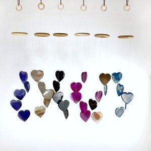 May include: A wind chime featuring multiple agate heart-shaped charms in various colors, including purple, pink, blue, black, and brown. The hearts are suspended from a wooden frame with strings.
